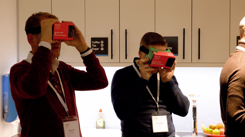 Two men wearing RNIB-branded virtual reality headsets in a kitchen area, focusing intently on the experience.
